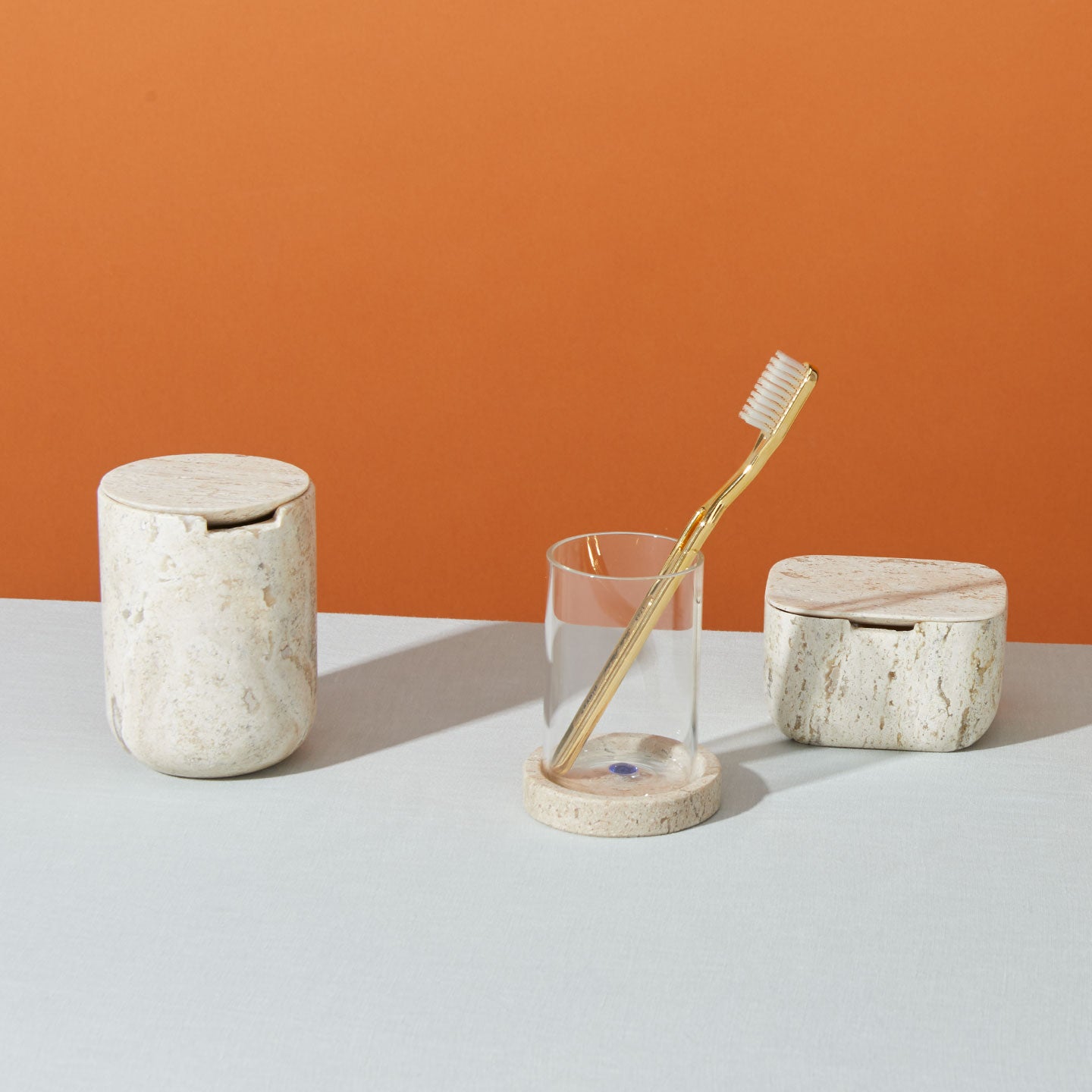 Three bathroom accessories are shown: a toothbrush holder with a transparent glass and a wooden toothbrush, a cylindrical container with a lid, and a rectangular container with a lid.