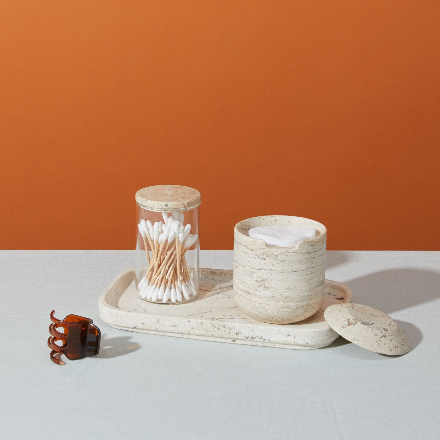 A still life of a beige stone tray holding a lidded jar with cotton pads, a lidded jar with cotton swabs, and a brown hair claw clip on a light blue surface, with an orange backdrop.