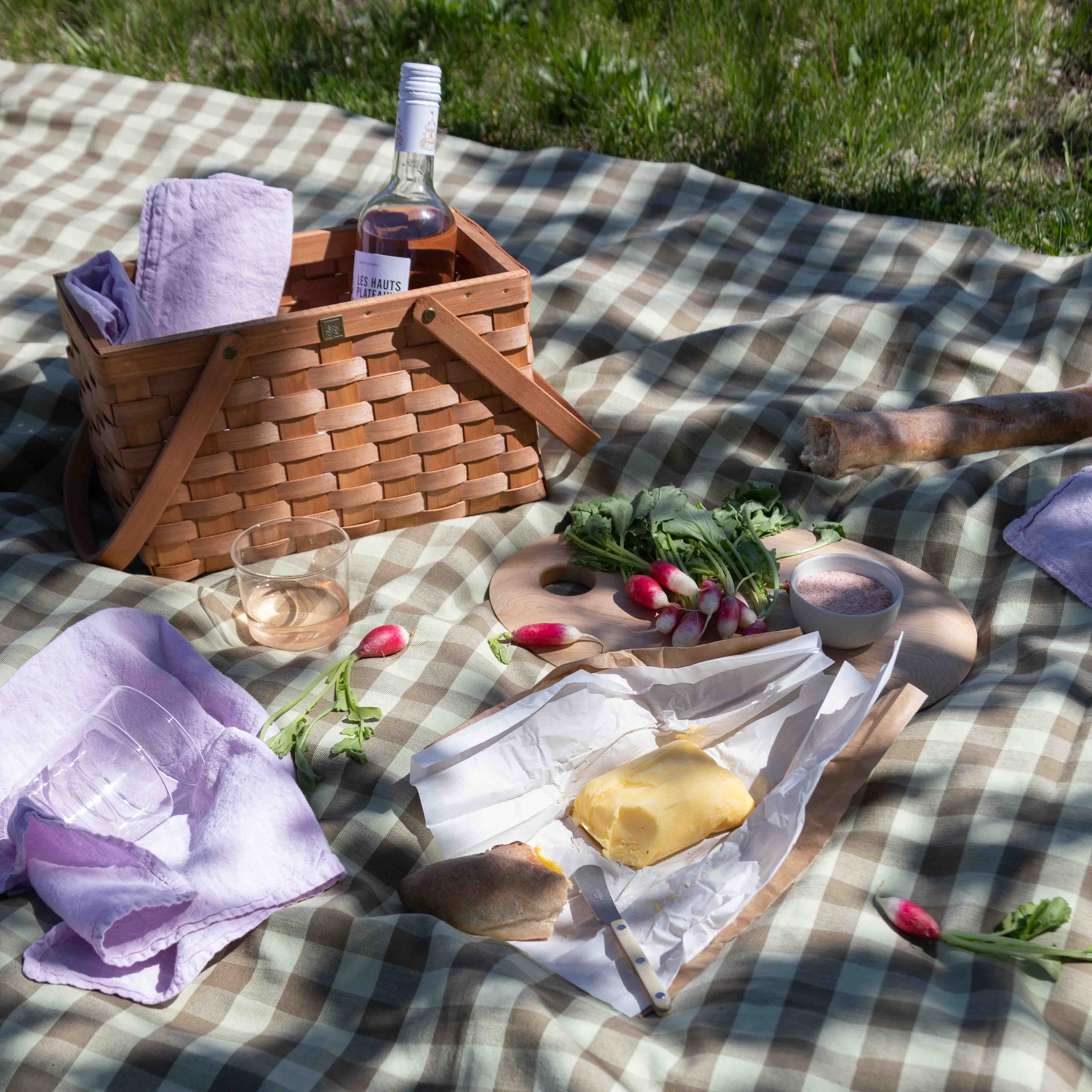 Picnic basket on gingham tablecloth and linen napkins with rose, butter, radishes and baguette.