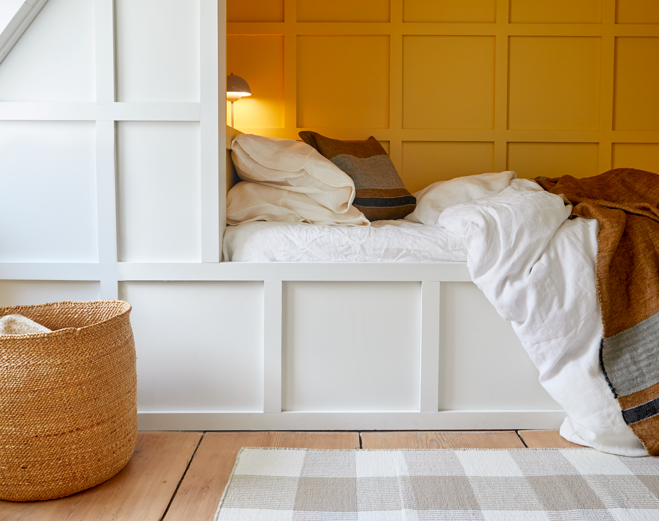 White built-in bed with bedding, a rug, and a basket in a room with white paneled walls.