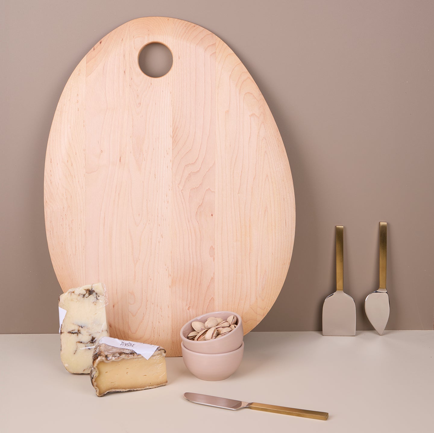 Maple pebble-shaped cutting board alongside an assortment of cheese, a set of three metal cheese knives, and a set of two Blush colored extra small bowls filled with nuts.
