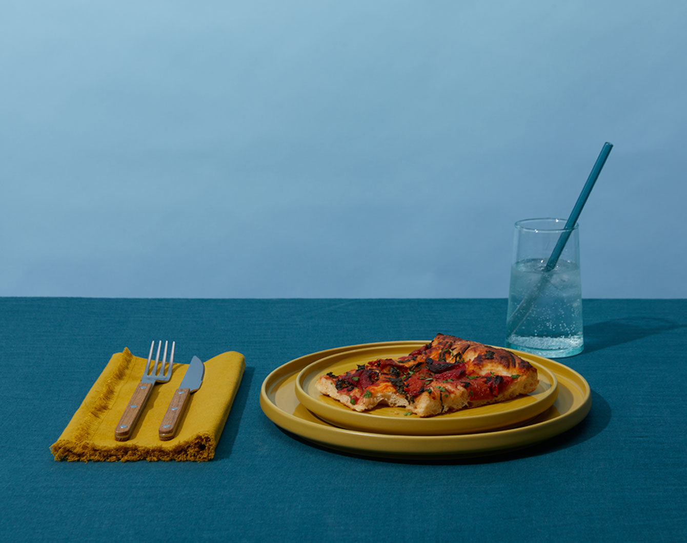 mustard colored dinnerware styled with a slice of pizza, a blue drinking glass and mustard linen napkin with teak flatware on a blue background