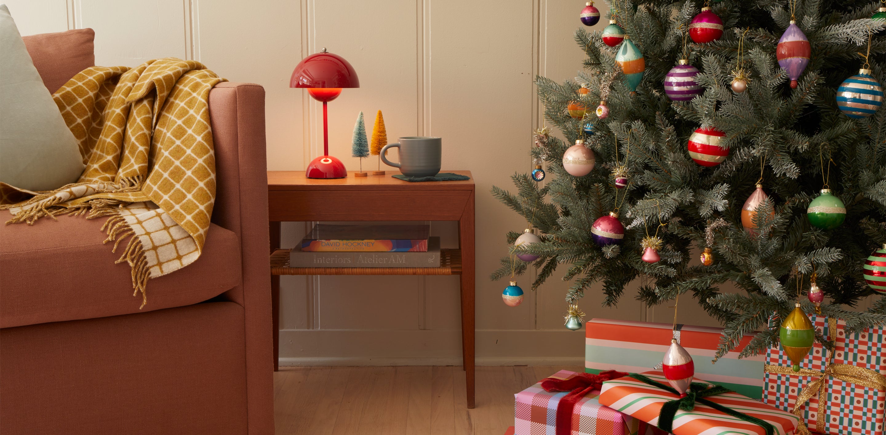 A decorated tree with wrapped gifts underneath, a sofa with a checked throw and with a side table with a lamp, mug and bottlebrush trees.