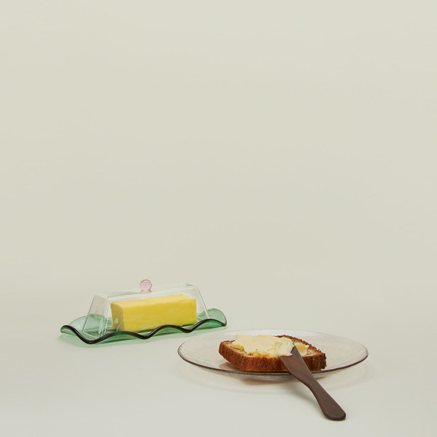 A glass butter dish with a stick of butter, a wood spreader, and a glass plate with buttered toast.