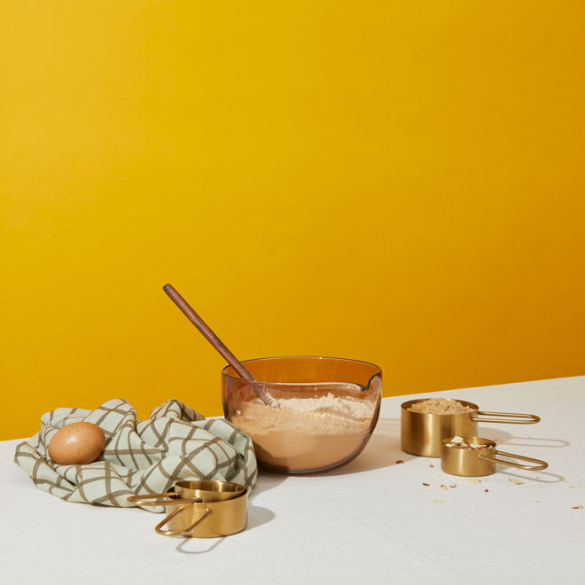 mixing bowl, measuring cups and kitchen towel styled with eggs and flour on yellow background