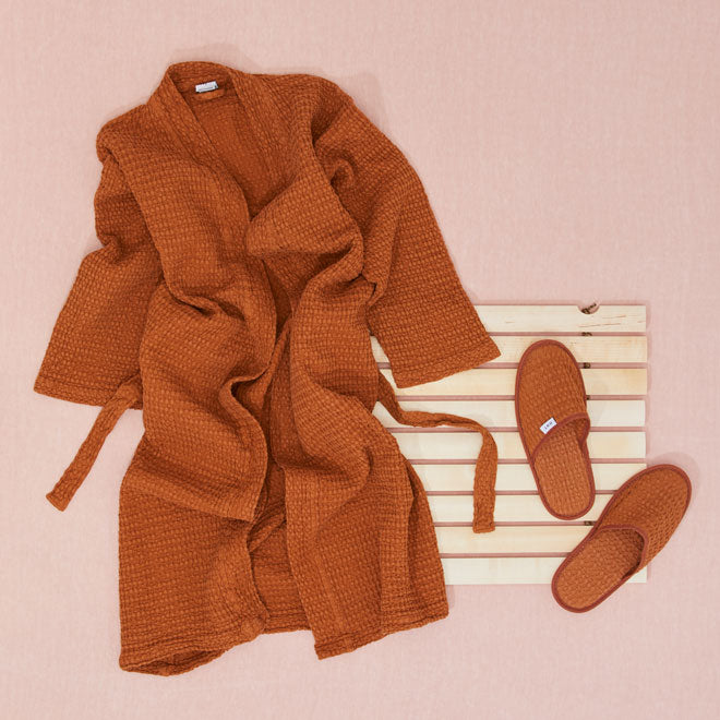 orange robe and slippers laying on wood slatted bath mat on pink background 