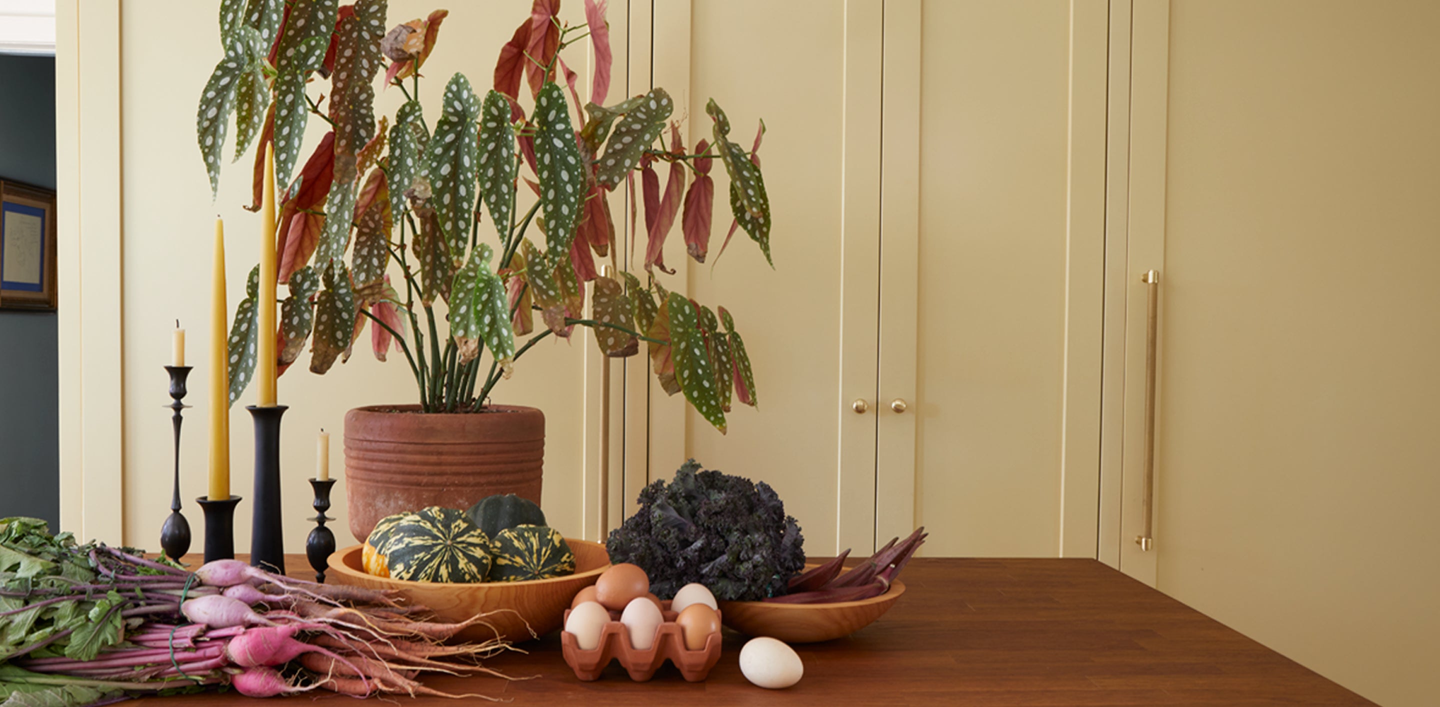 A table with a large potted alocacia plant, taper candles, wooden bowls containing various vegetables,a bundle of radishes and an egg holder