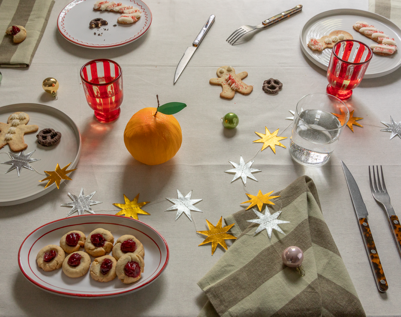 Table setting with plates, glasses, napkins, holiday decorations, and cookies scattered on an ivory colored tablecloth