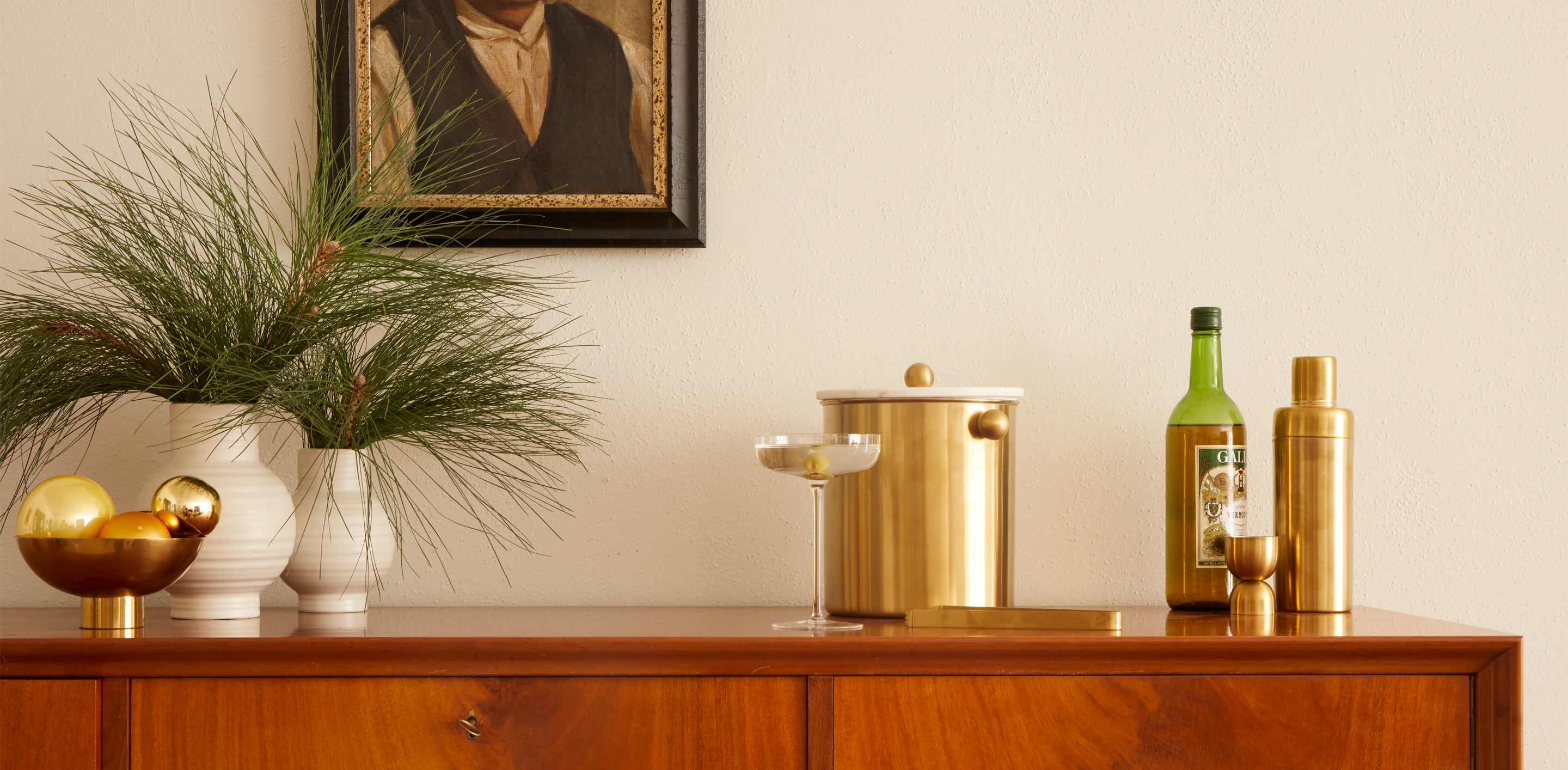 A simple bar with artwork in the backgrouns with an ice bucket and barware, two vases hold cut foliage and a brass footed bowl holds holiday ornaments