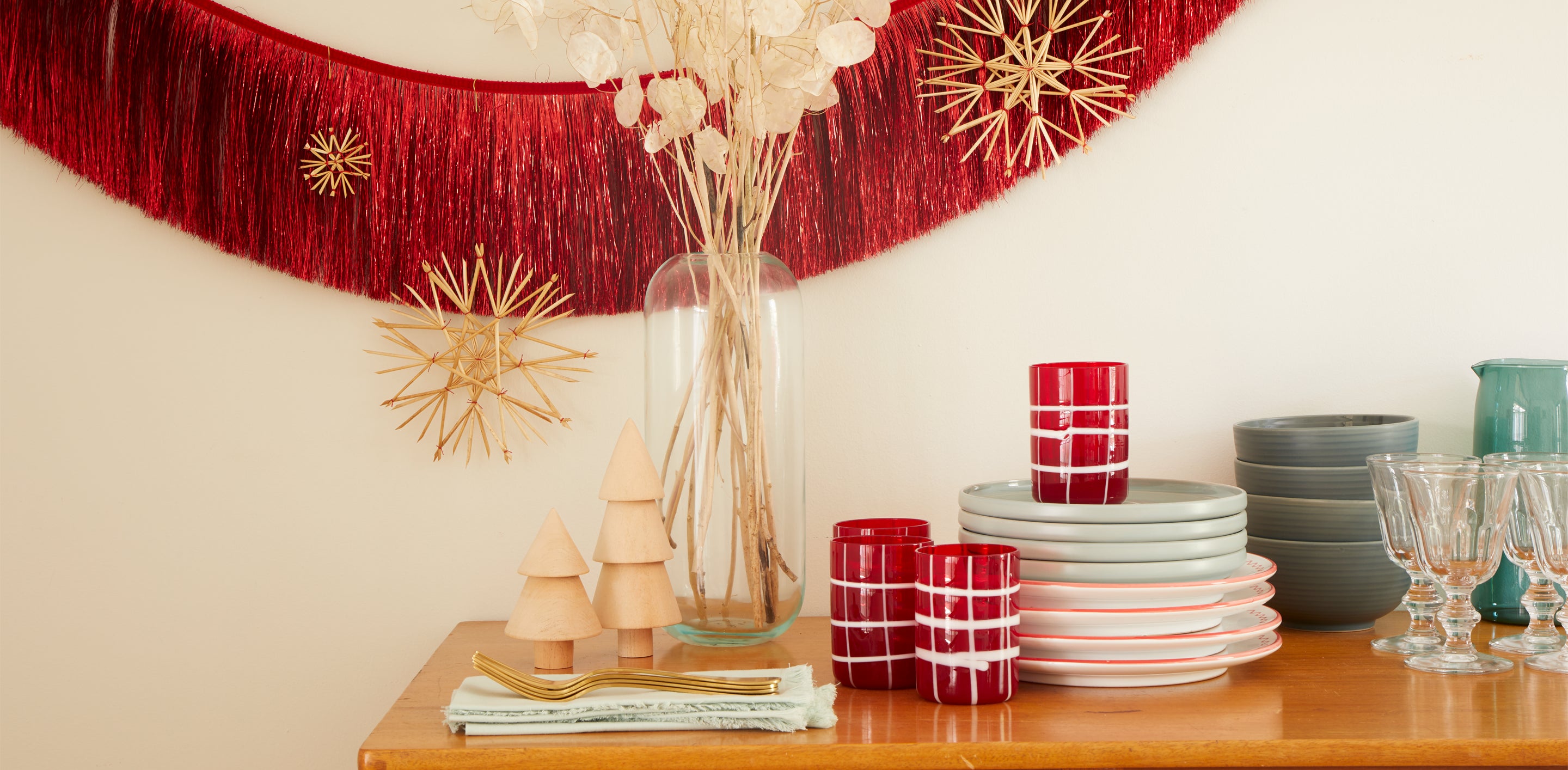 Table with festive dinnerware, glassware, a vase with dried florals and turned wooden trees, a red garland hanging in background with straw ornaments in various sizes. 