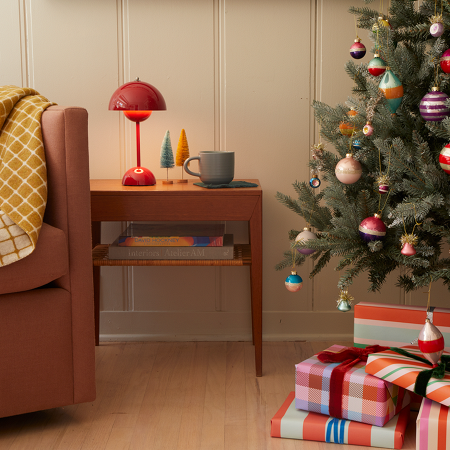A decorated tree with wrapped gifts underneath sits nest to a sofa with a checked throw and an end table with a lamp, bottlebrush trees and a mug