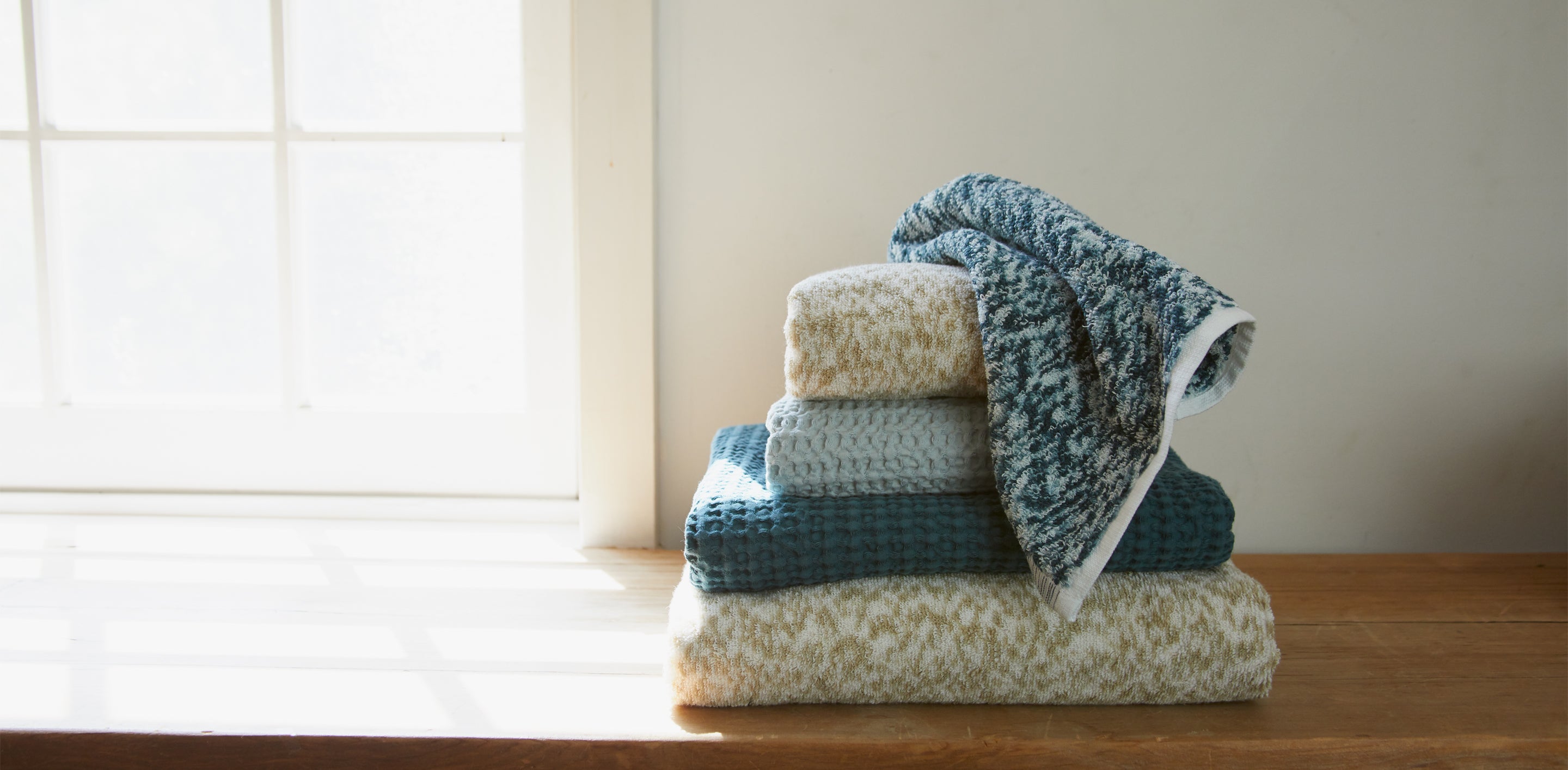 A stack of folded towels in various patterns and colors sits on a wooden surface near a window with sunlight streaming through.