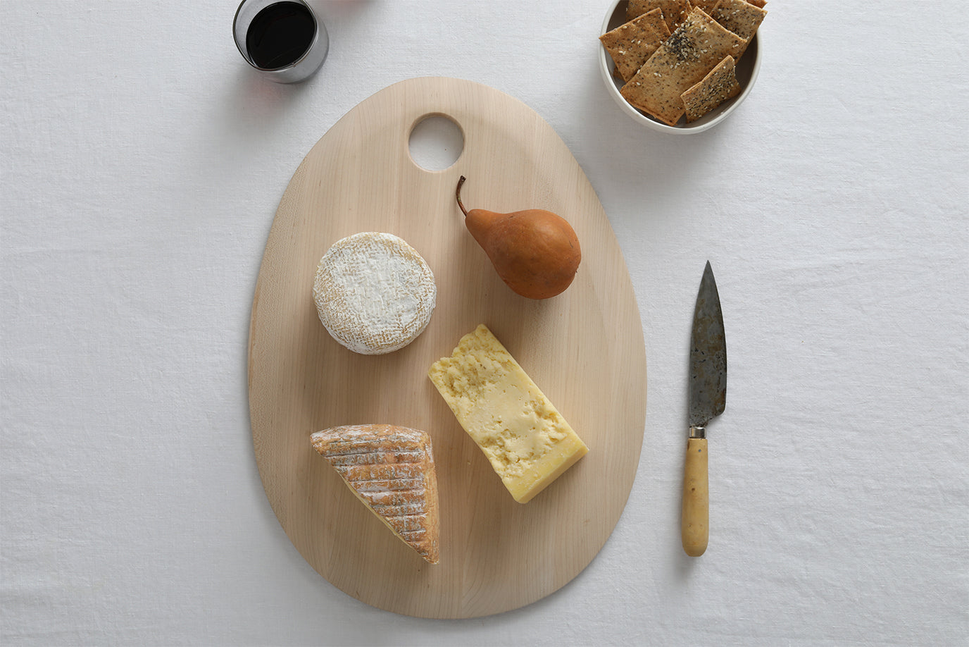 A cutting board and knife with a variety of cheeses, a pear, a bowl of crackers and a glass of red wine.