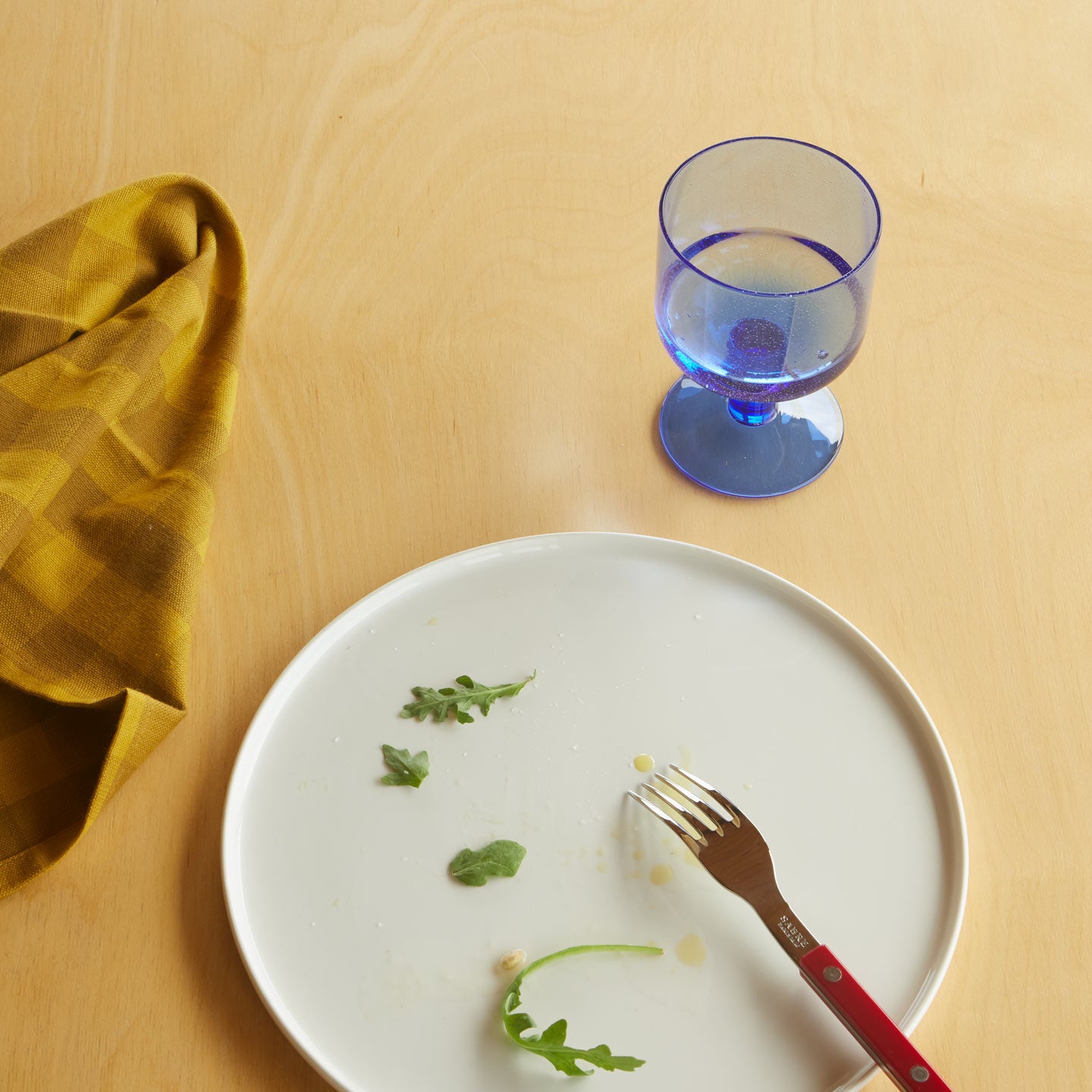 Overhead of place setting on wood tabletop with essential Gingham Napkin in Mustard, Blue Seeded Glass, and Simple Porcelain Plate with red fork.