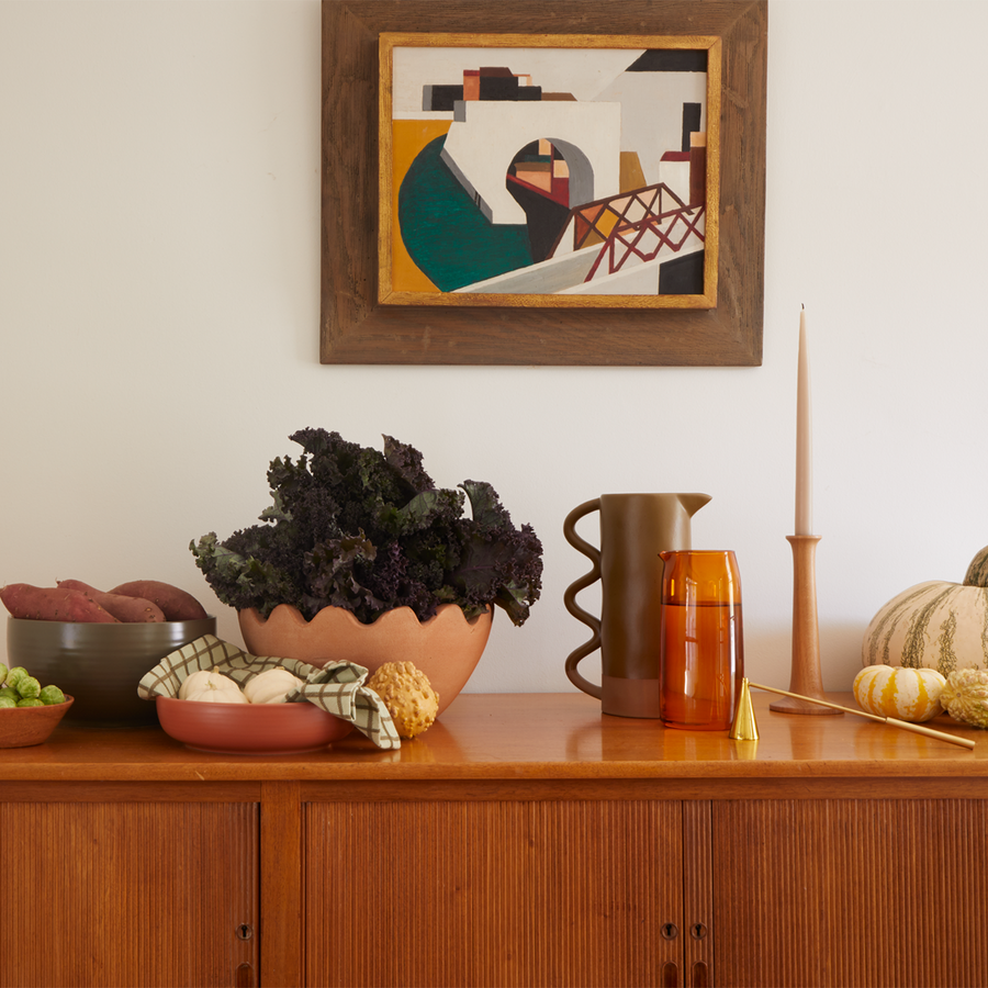 A sideboard with large serving bowls of seasonal vegetables sits next to a ceramic and a glass pitcher with a candle douter and pumpkins. one the wall hangs a piece of modern artwork