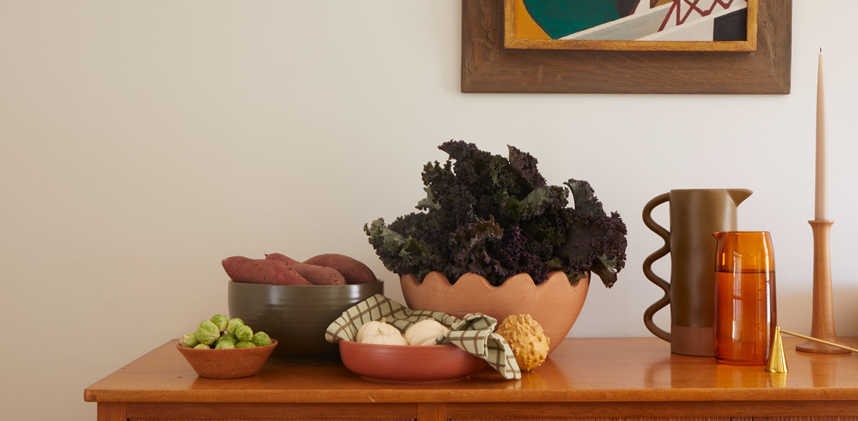 A dining table with serving bowls of various vegetables, to the right sits a clay and glass pitchers, a candle douter, taper candle, and artwork hangs in the background