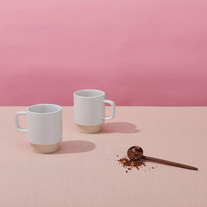 Two mugs and a scoop for coffee and tea on a pink tabletop.