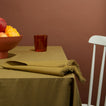 Table setting with a bowl of fruit, glass, and folded napkin on an olive tablecloth against a brown wall.