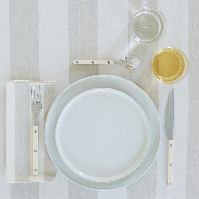 A place setting of essential dinnerware, essential glassware and flatware arranged on a striped tablecloth with striped napkin.
