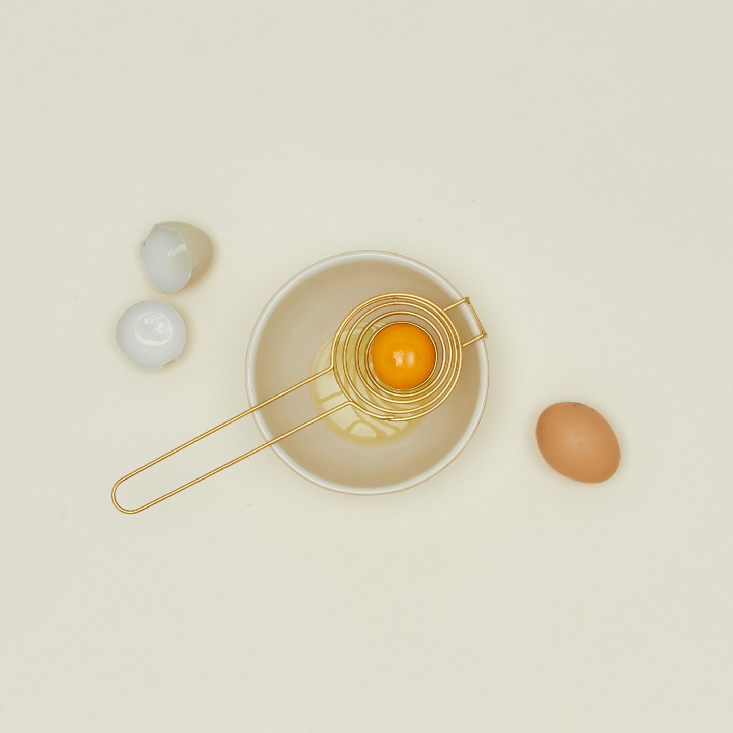 Brass swirl egg separator with egg yolk over bowl and egg shells on ivory background.