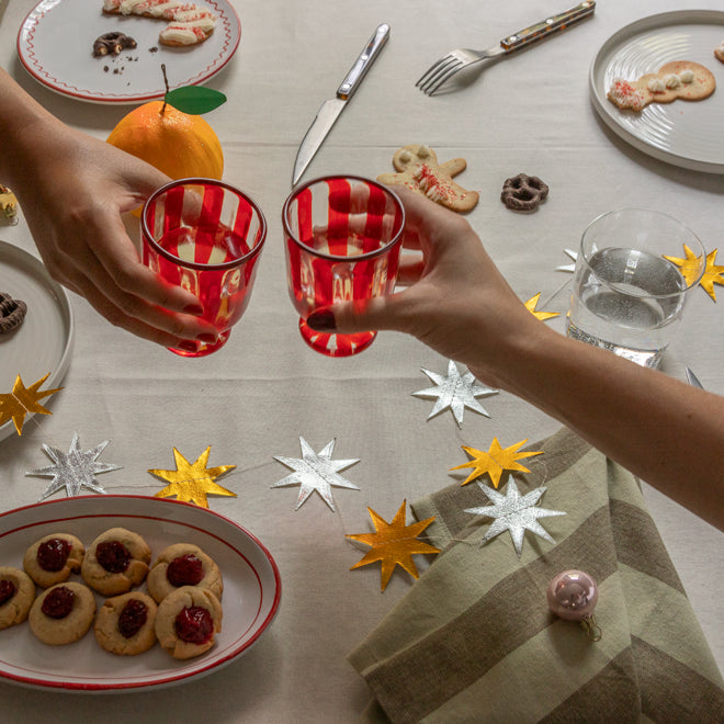 Two hands clinking red glasses on a festive table with cookies and decorations.