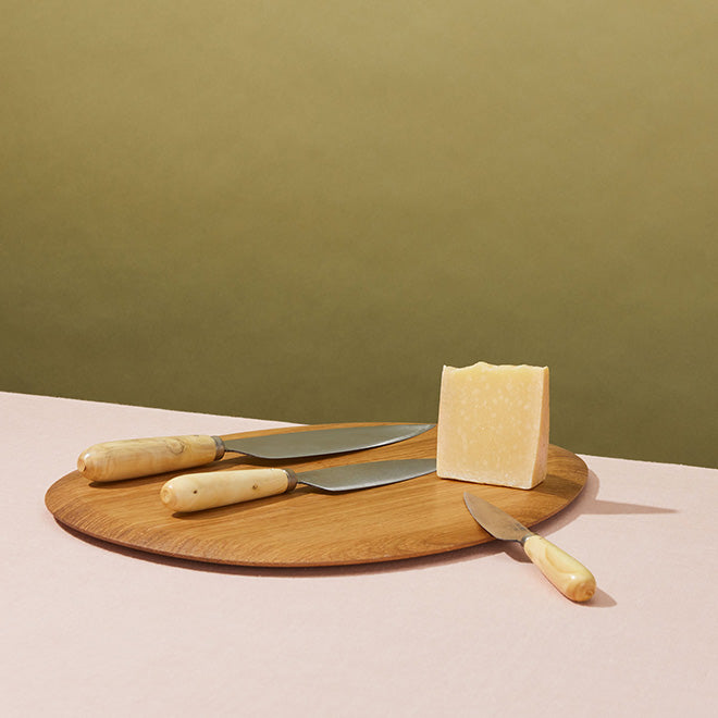 A kitchen cutting board and knives with cheese on a pink tabletop.