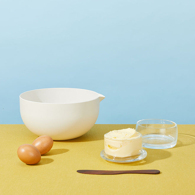 Kitchen mixing bowl, butter keeper and butter knife with eggs on yellow tabletop.